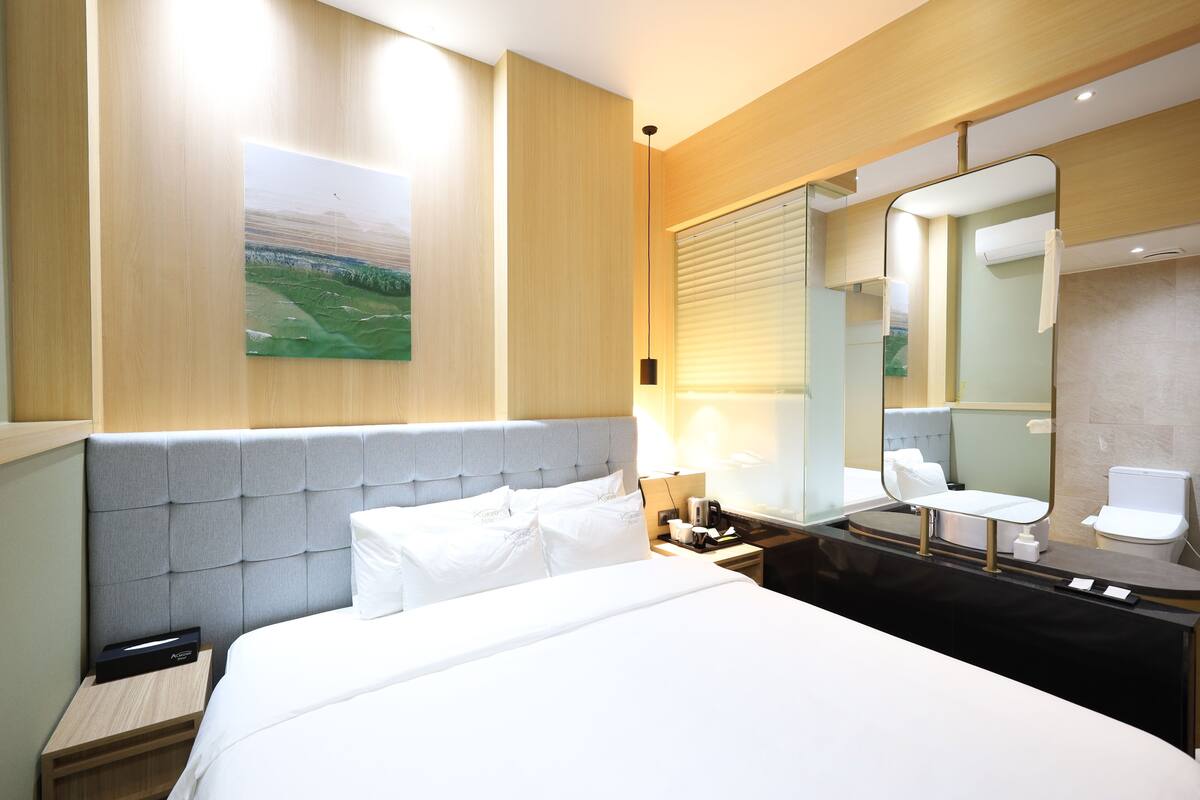 A hotel room with a bed, a nightstand, a mirror, and a window with blinds.