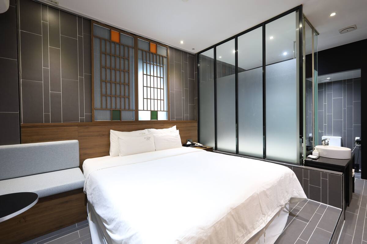 A modern hotel room with a large bed, a bedside table, a sofa, a large window with a view, and a separate bathroom area.