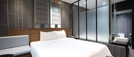 A modern hotel room with a large bed, a bedside table, a sofa, a large window with a view, and a separate bathroom area.
