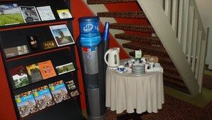 Una escalera con un suelo de alfombra estampada en rojo y marrón, una estantería negra con varios objetos, una mesa con mantel blanco y juego de té, y una botella de agua azul.