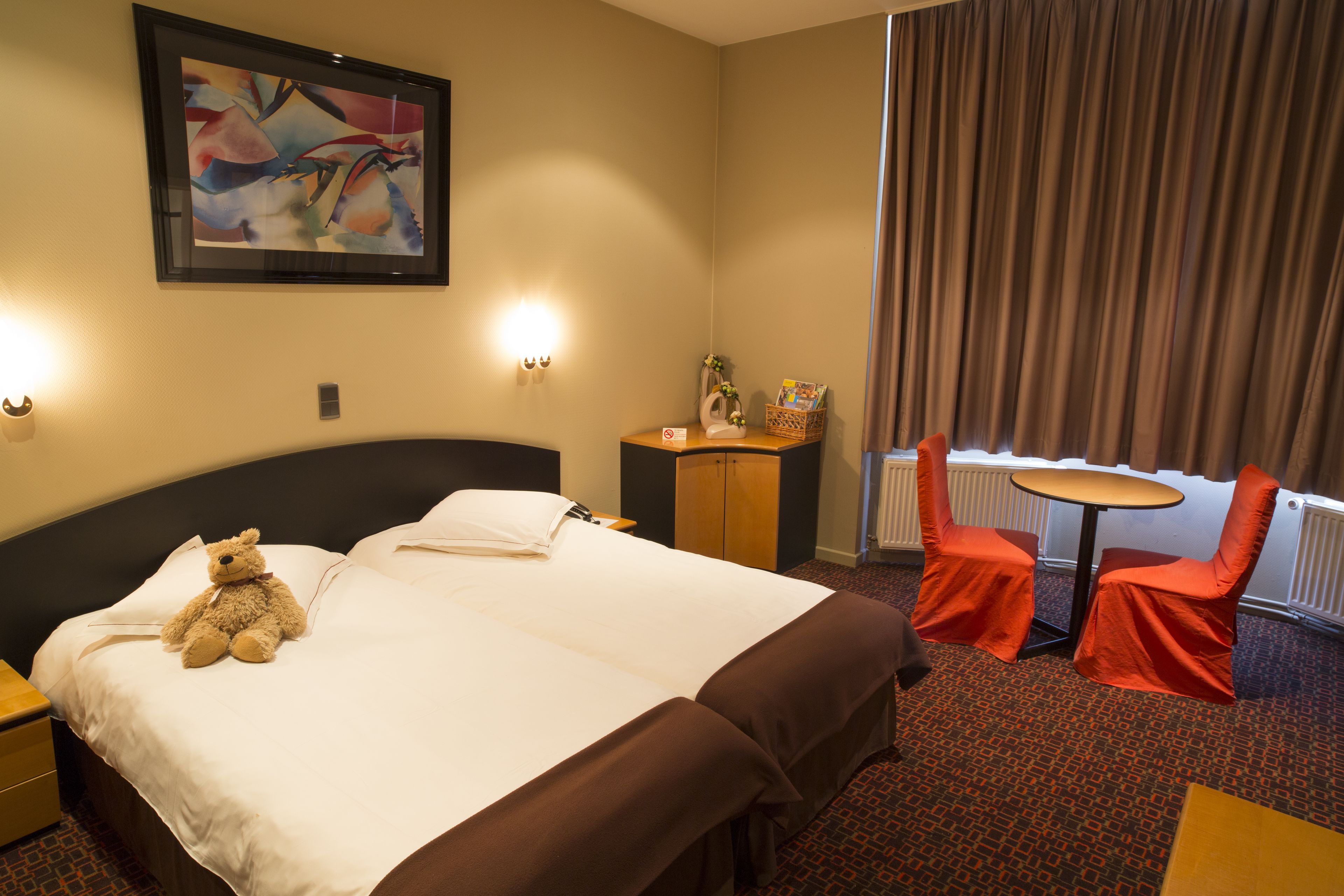 Hotelkamer met een groot bed, nachtkastje, stoel, klein tafeltje en een raam met gordijnen.