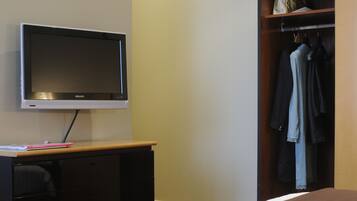 A hotel room with a flat-screen TV on a wooden desk, a wardrobe with hanging clothes, and a bed with a brown blanket.