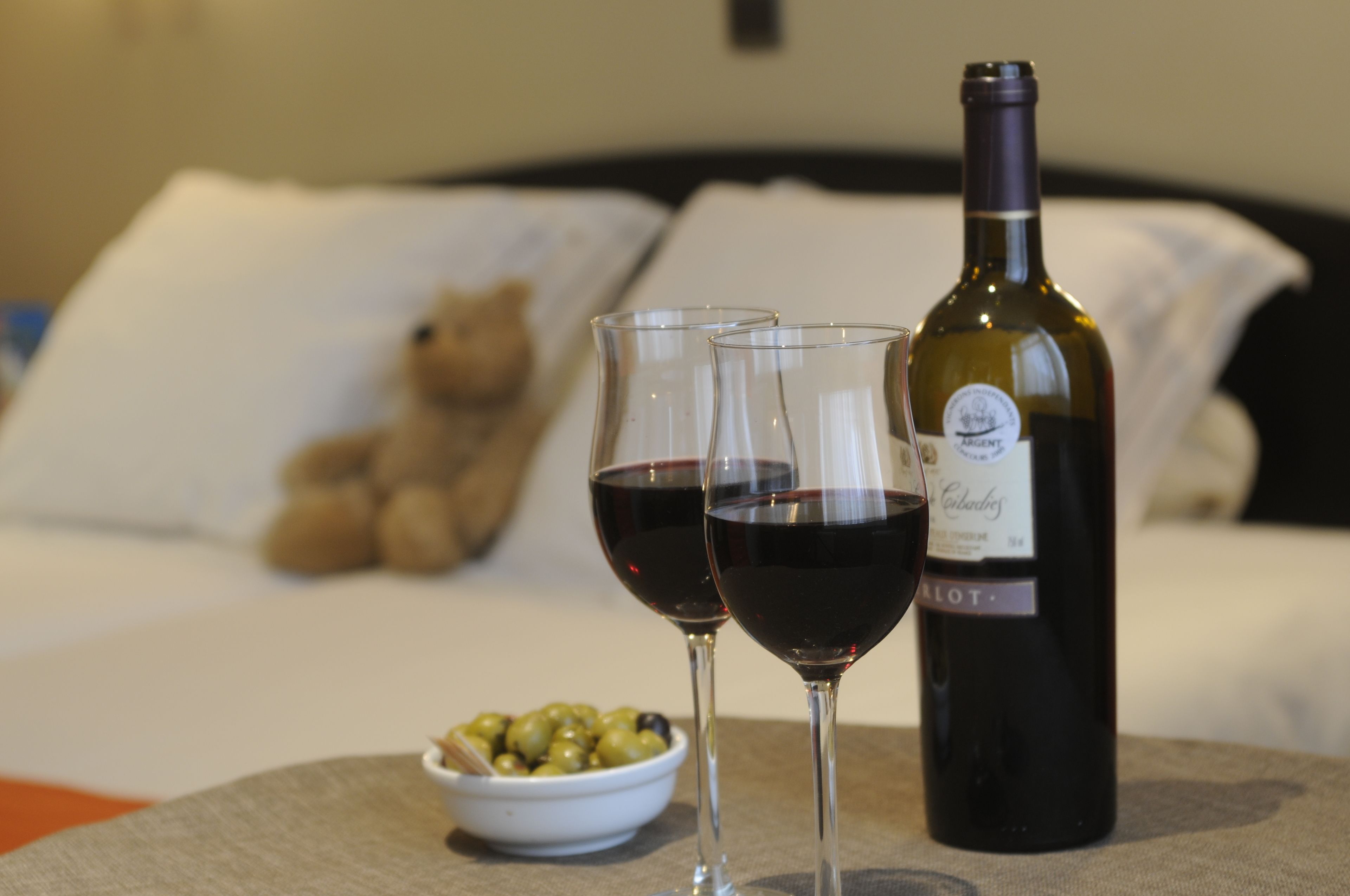 A bottle of wine, two glasses of red wine, and a bowl of olives on a table in a hotel room.