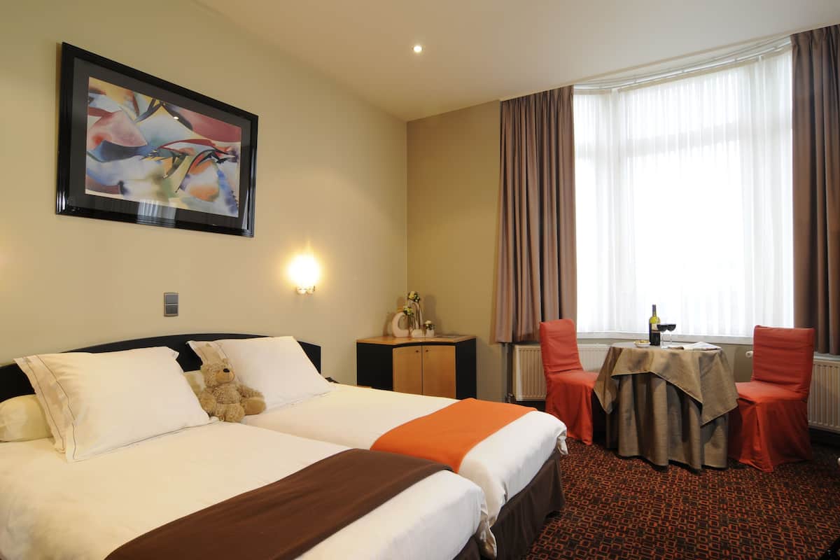 A hotel room with two beds, a table with chairs, a painting on the wall, and a window with curtains.
