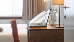 Habitación de hotel con cama, escritorio con una laptop, lámpara y ventana con cortinas.