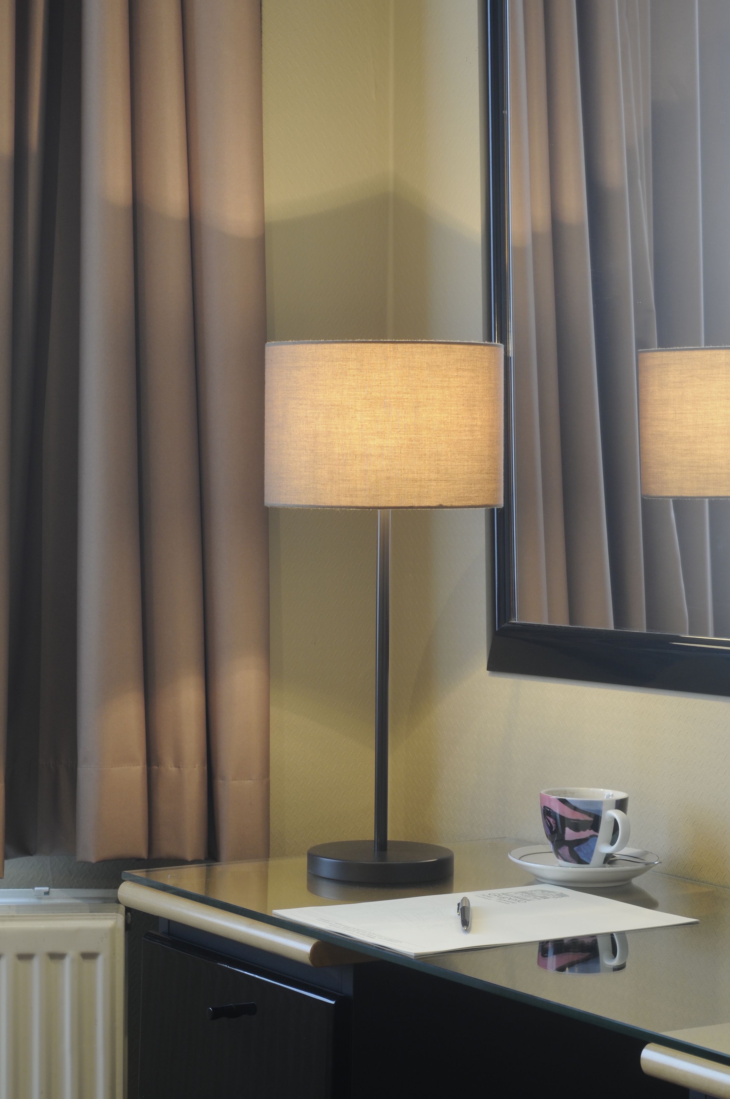 A hotel room with a desk, a lamp, a cup, and a mirror.