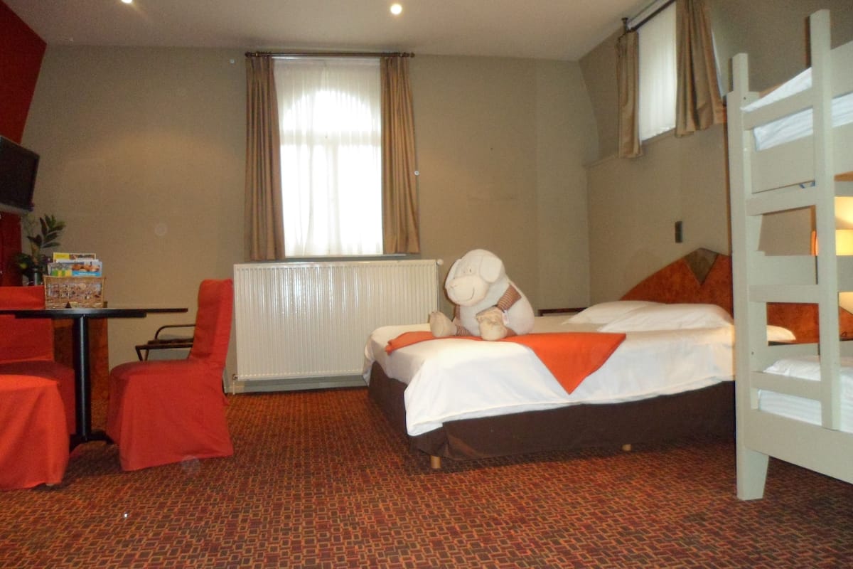 A hotel room with a bed, a red chair, a table with a board game, a radiator, and a window with curtains.