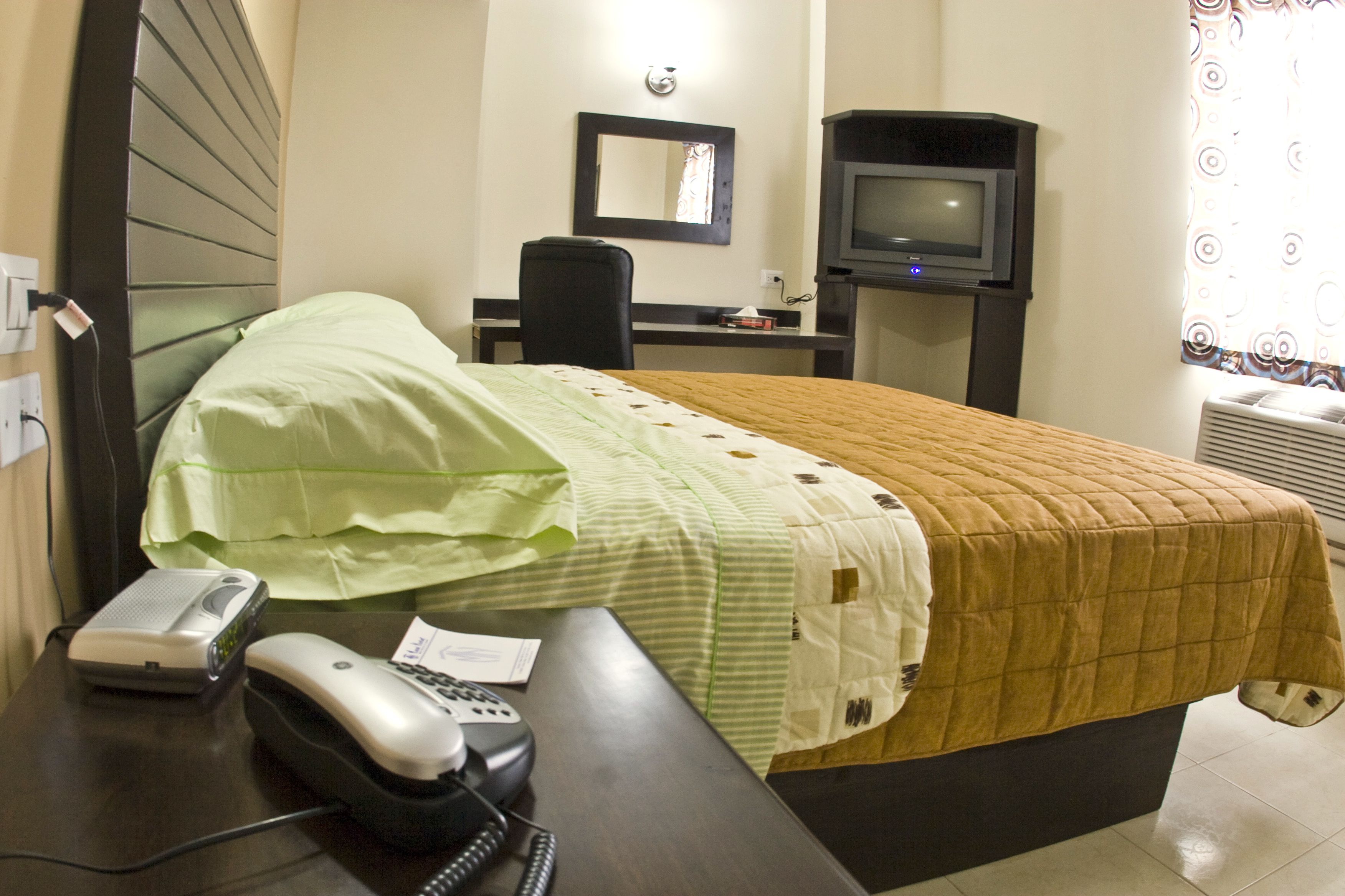 A hotel room with a bed, a desk with a telephone and a speaker, a chair, a television, and a window with a curtain.