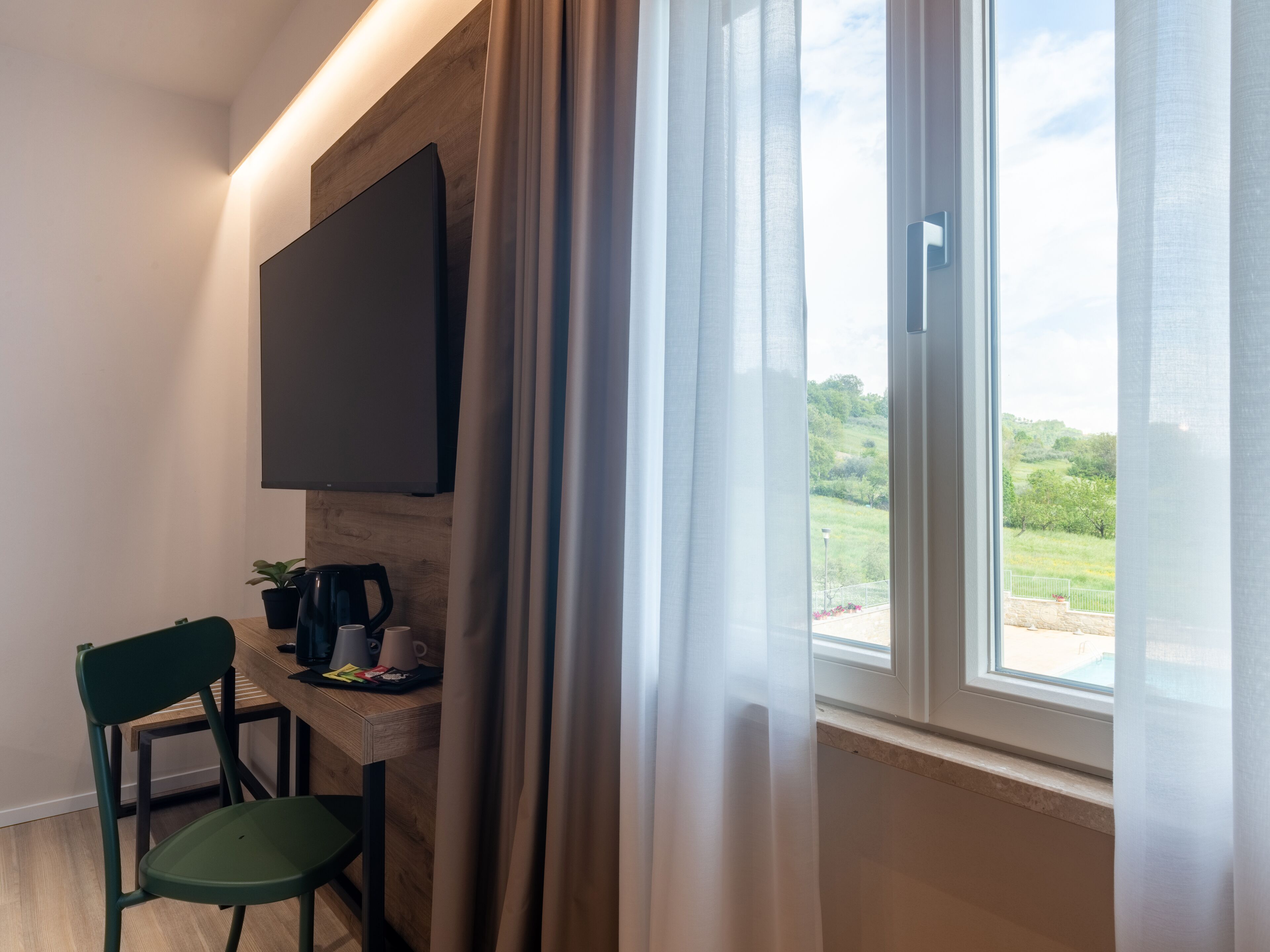 A room with a flat-screen TV mounted on a wooden wall, a table with a coffee maker, cups, and a plant, and a chair.