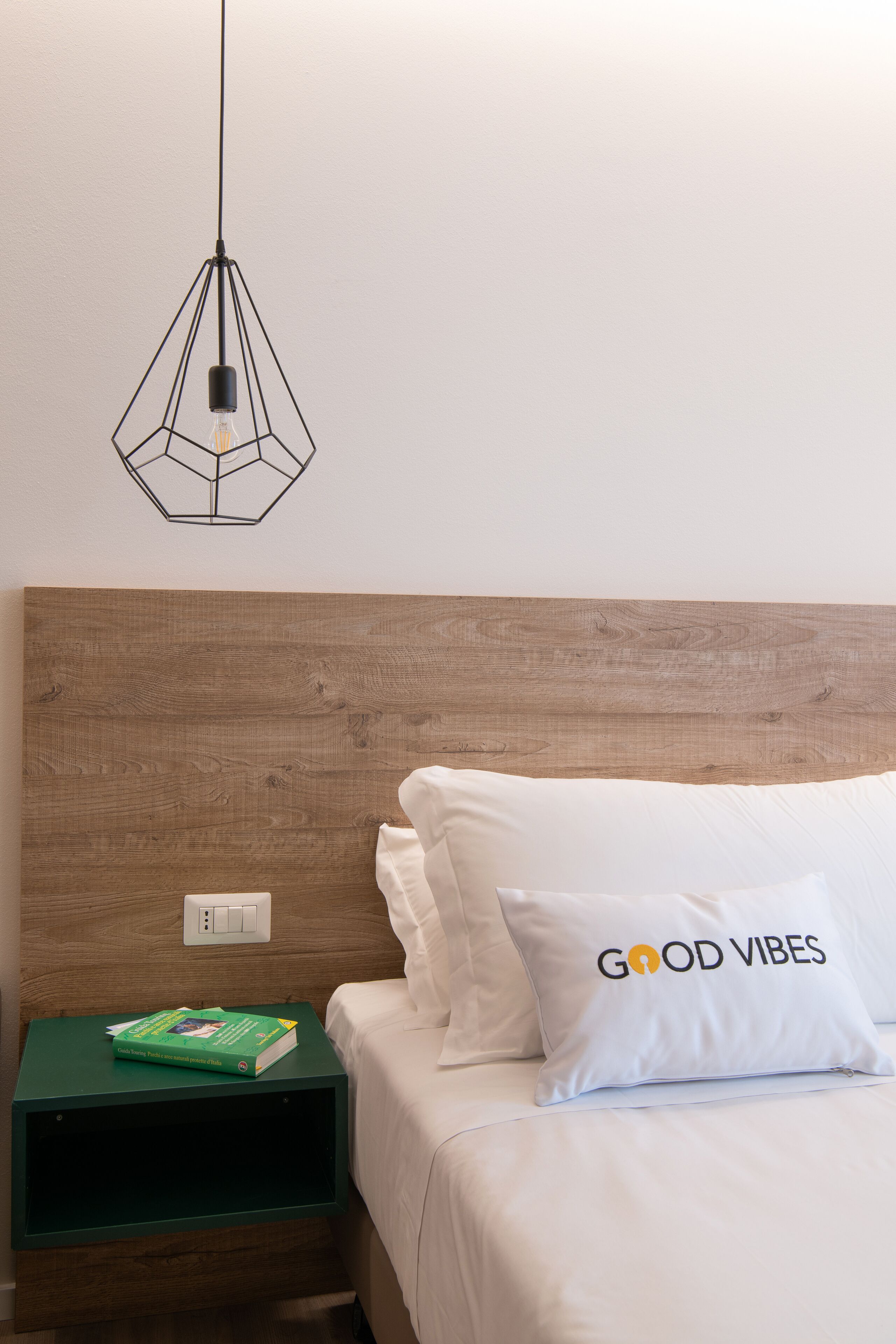 A neatly made bed with a pillow labeled "GOOD VIBES" and a green nightstand with a book on it.