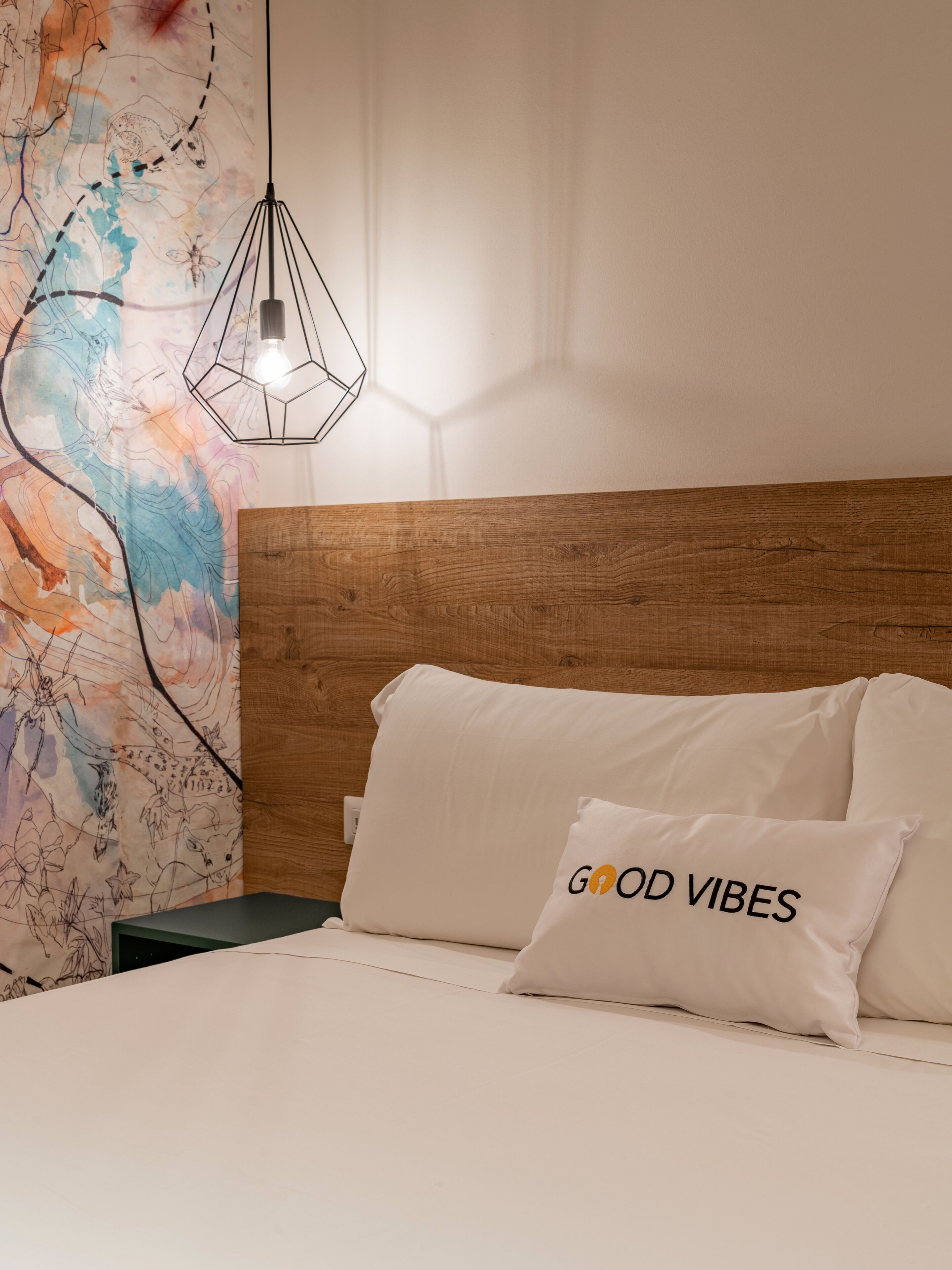 Un dormitorio con cabecera de madera, un mural abstracto y colorido en la pared, una lámpara colgante moderna y un cojín con el texto "GOOD VIBES".