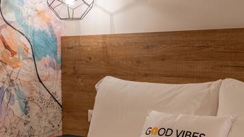 Un dormitorio con cabecera de madera, un mural abstracto y colorido en la pared, una lámpara colgante moderna y un cojín con el texto "GOOD VIBES".