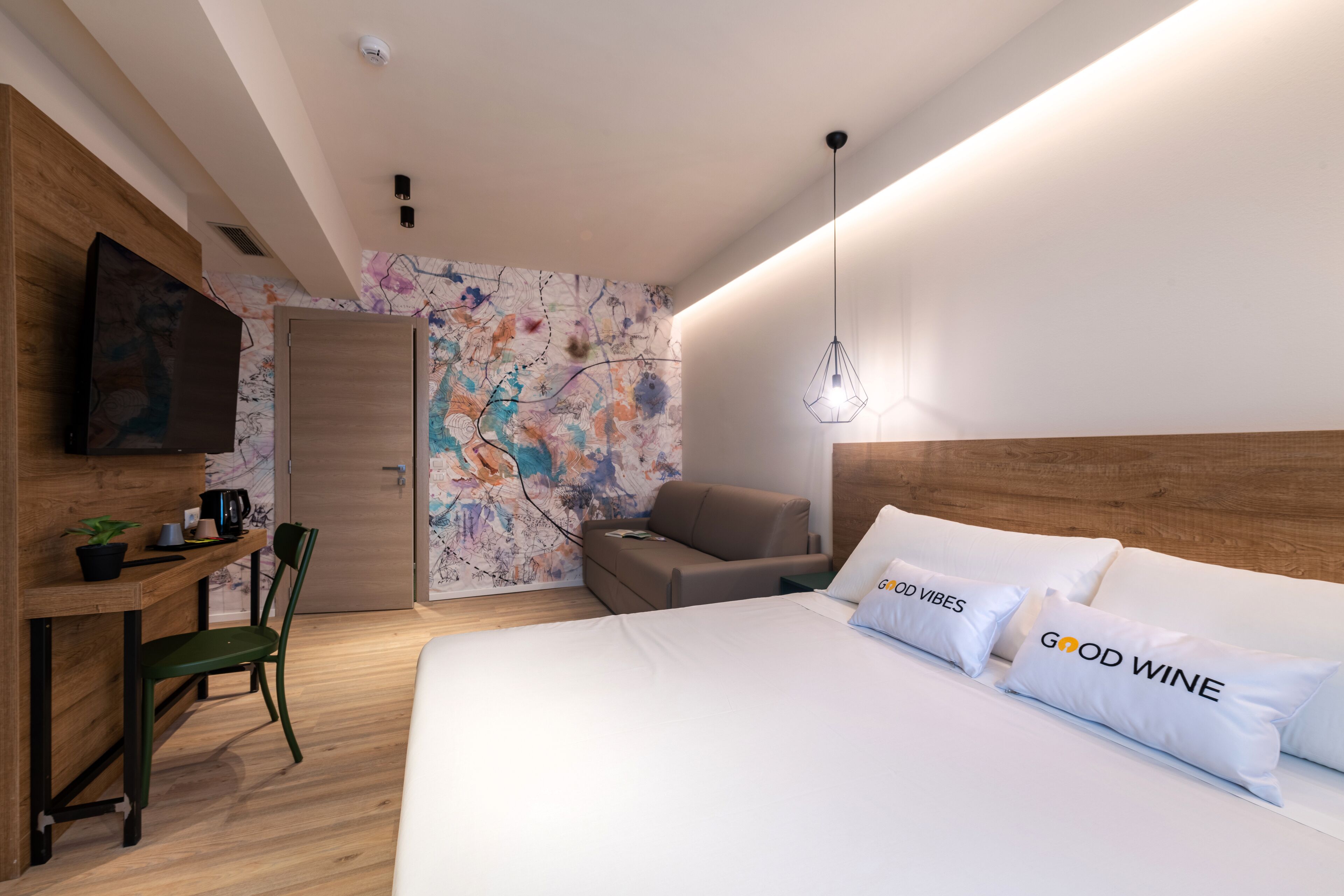 A modern hotel room with a wooden headboard, a bed with white bedding and pillows, a desk with a chair, a television, and a colorful abstract wall.