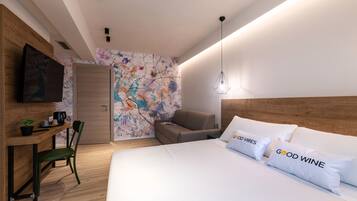 A modern hotel room with a wooden headboard, a bed with white bedding and pillows, a desk with a chair, a television, and a colorful abstract wall.