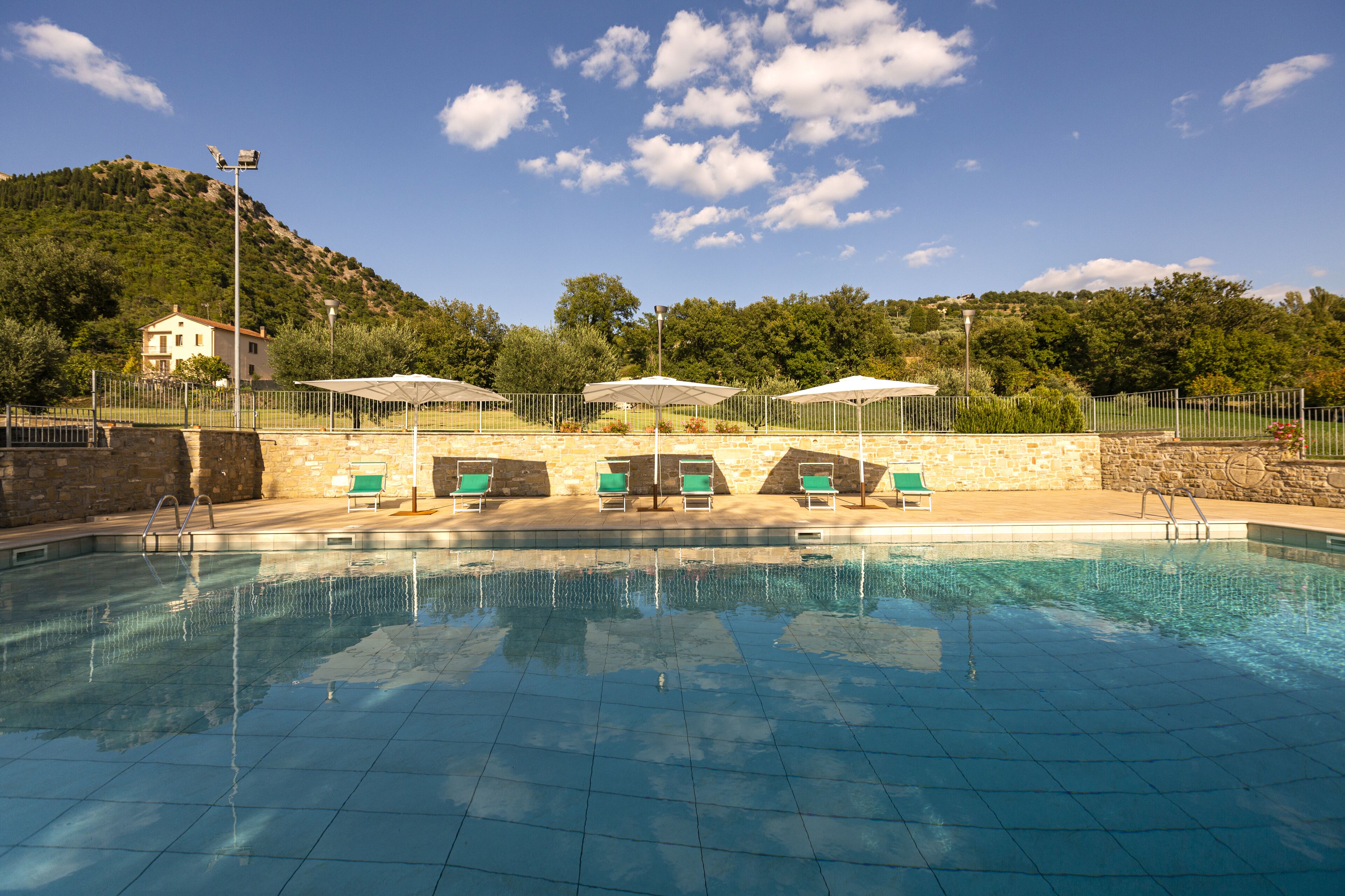 Una piscina con agua azul cristalina, rodeada por un muro de piedra y una valla de metal. Varias sillas de descanso y sombrillas se encuentran bajo una zona techada.