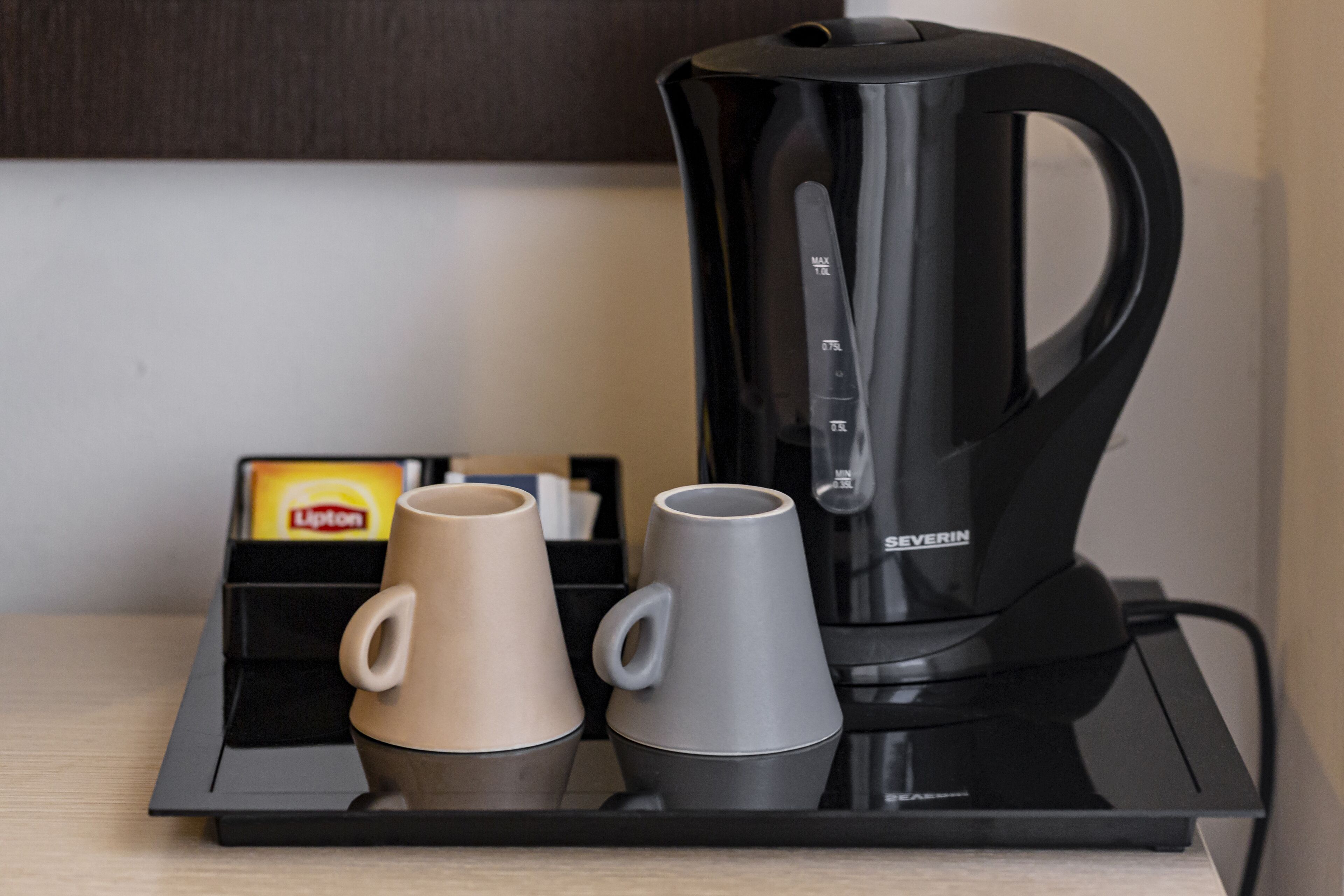 A black coffee maker on a tray with two cups, one beige and one gray, and a Lipton tea box in the background.