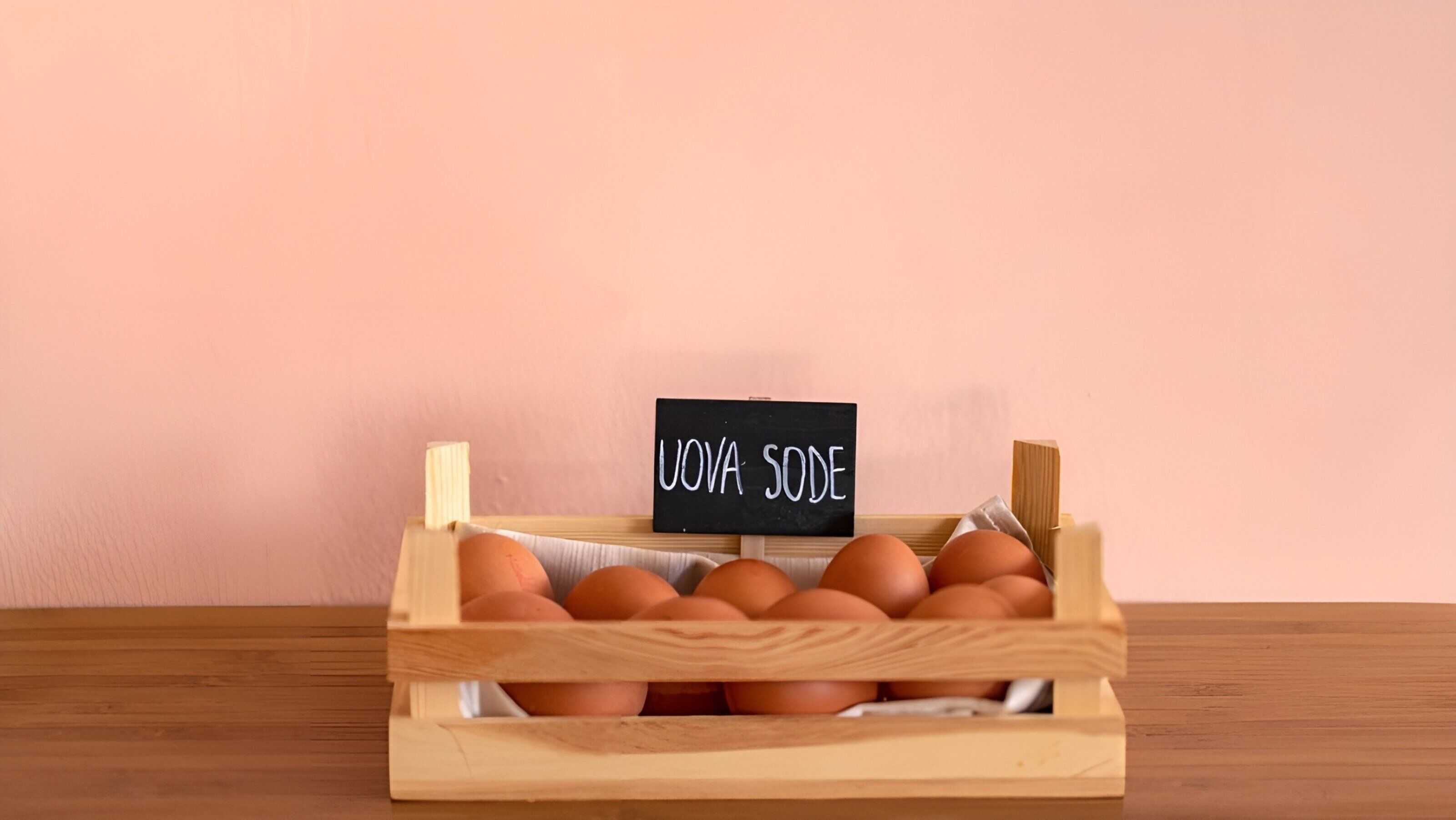 A wooden crate with eggs labeled "UOVA SODE" on a wooden table against a pink wall.