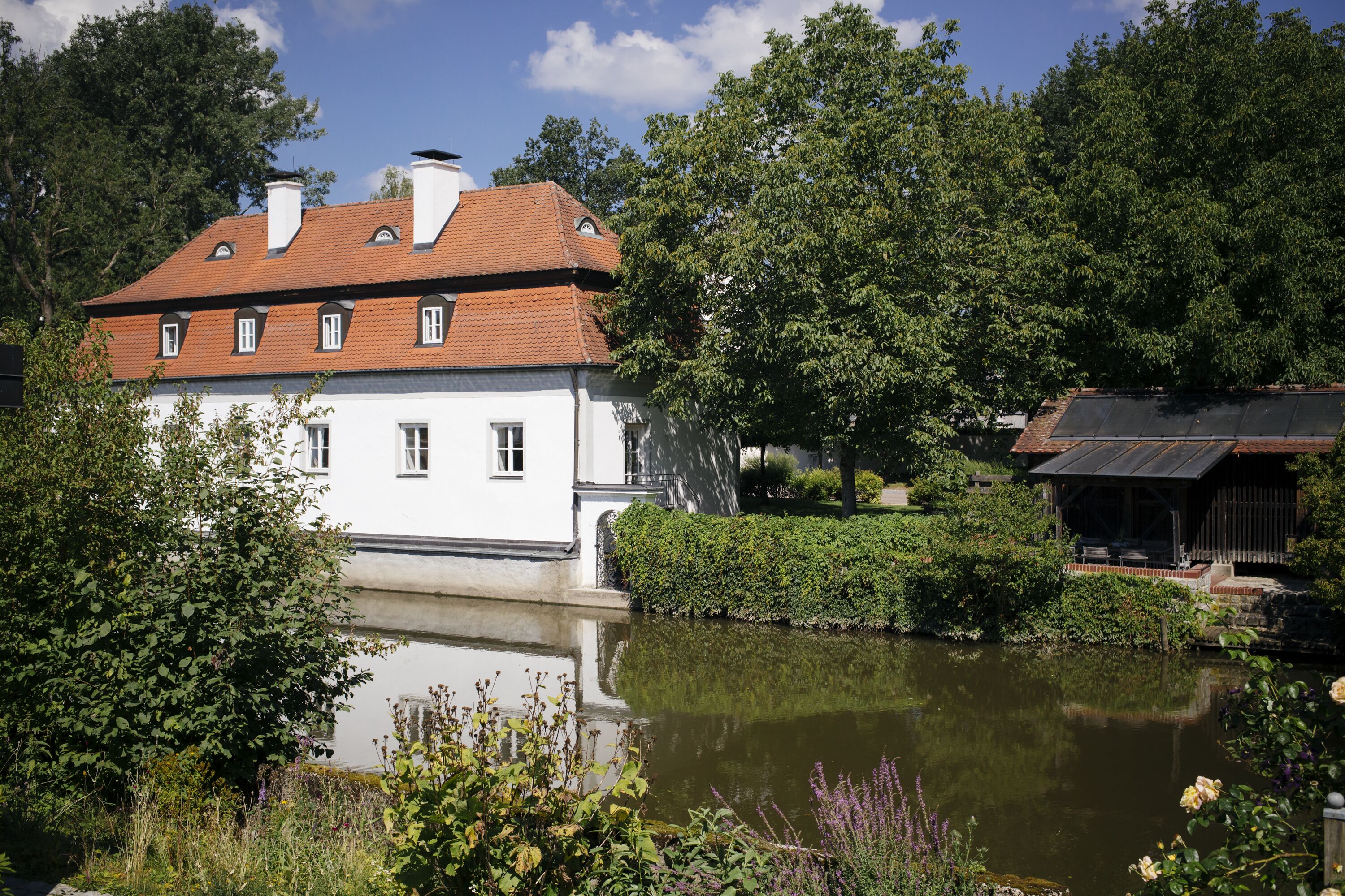 A white building with a red roof and multiple windows, situated next to a calm body of water, surrounded by lush greenery and trees.