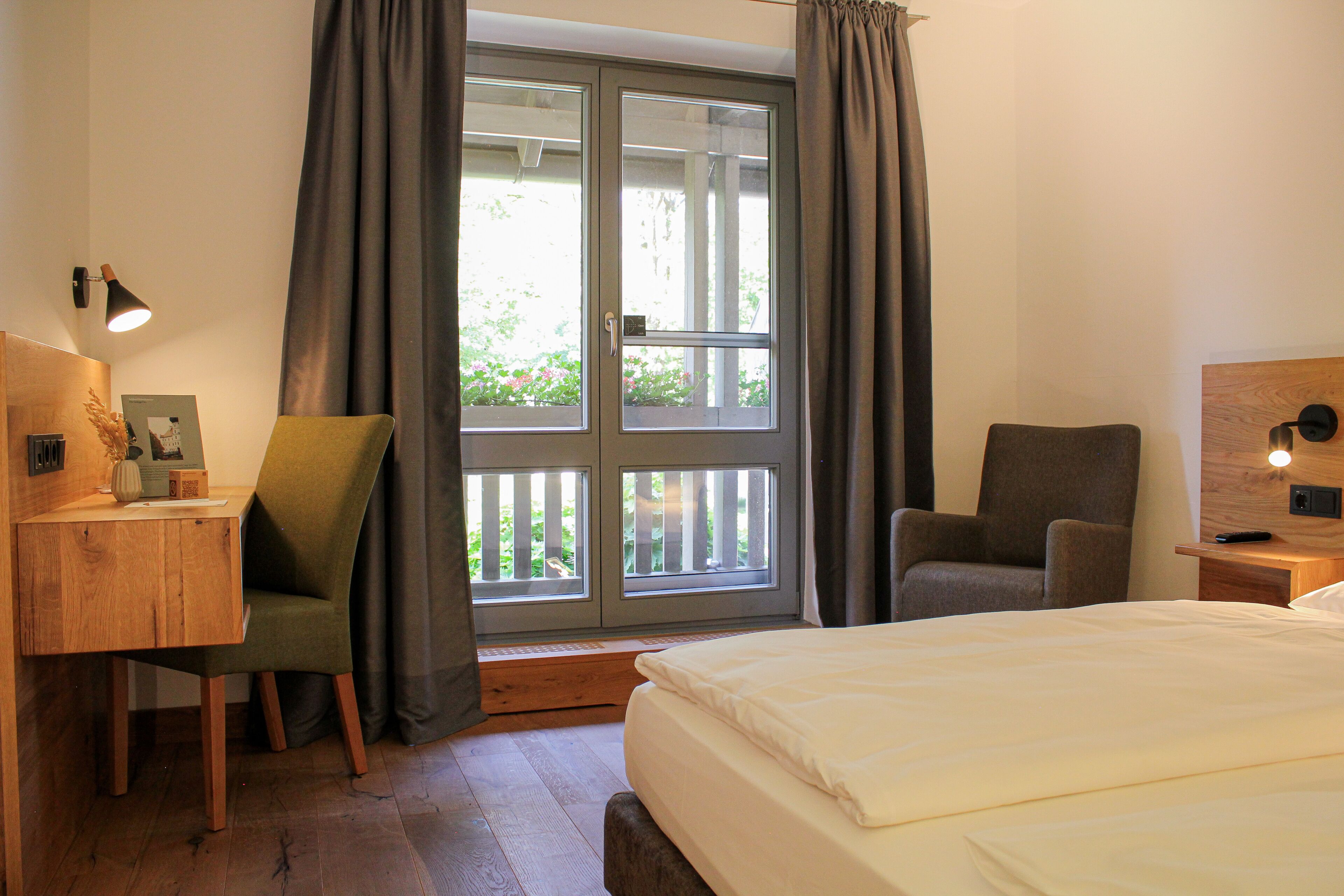 A bedroom with a bed, two chairs, a desk, a window with curtains, and a nightstand with a lamp.