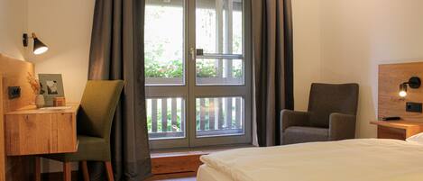 A bedroom with a bed, two chairs, a desk, a window with curtains, and a nightstand with a lamp.