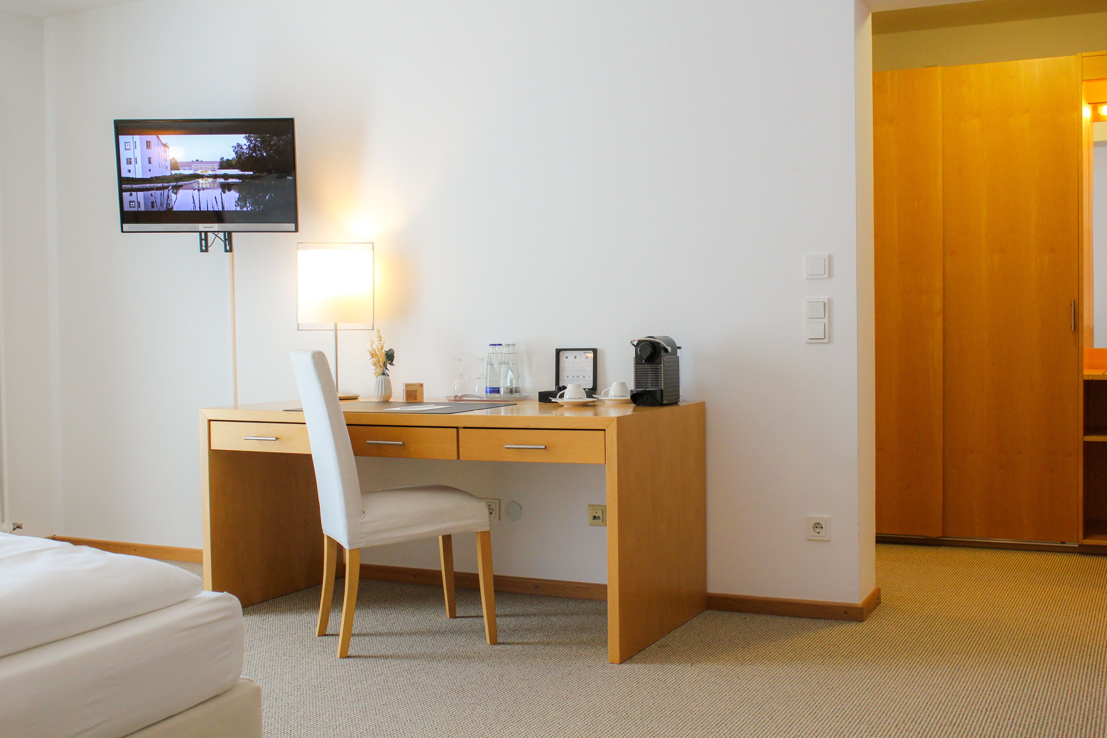 A hotel room with a wooden desk, a chair, a television mounted on the wall, a bedside table with a lamp, and a wardrobe.