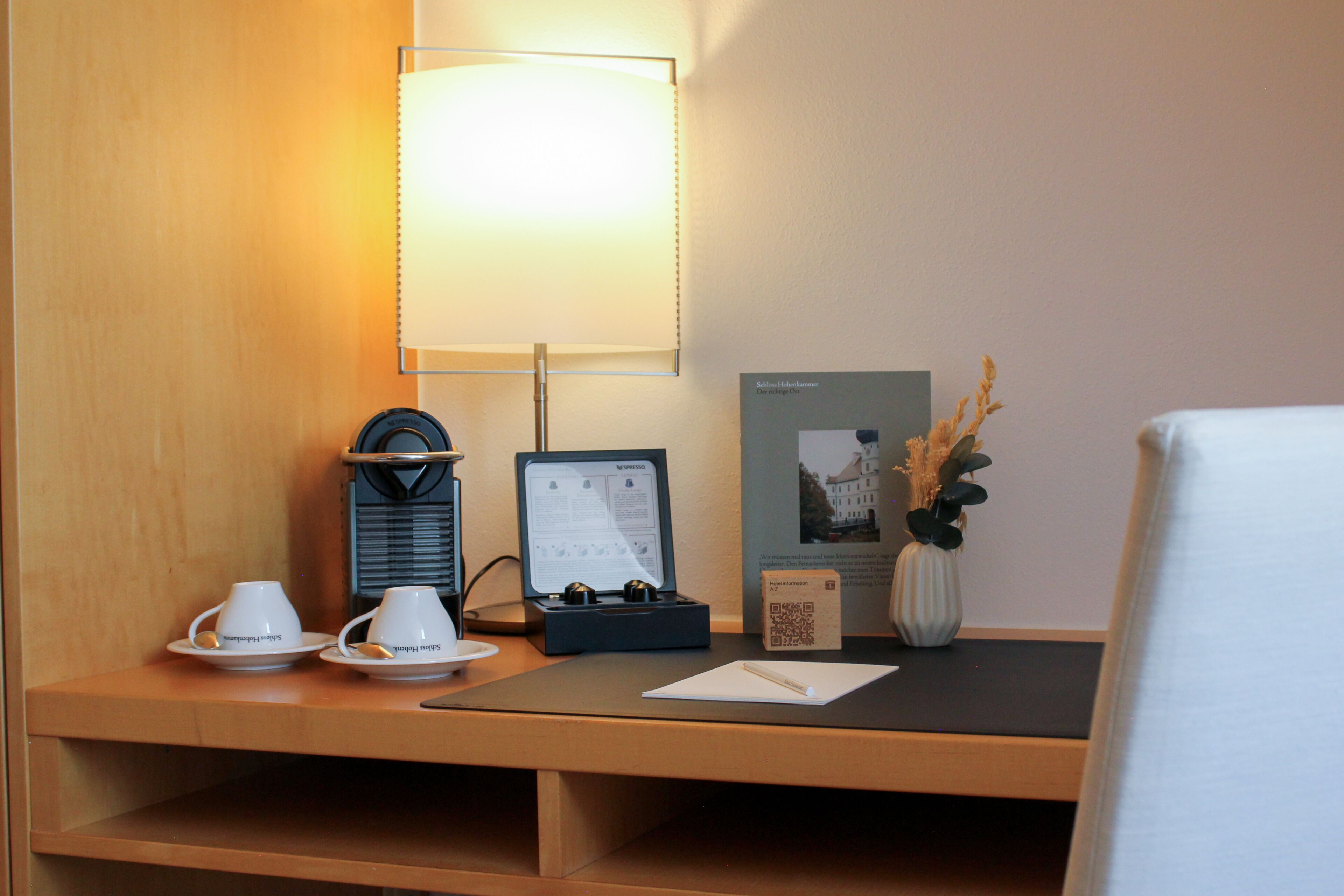 Un mostrador de hotel con una lámpara, dos tazas, un juego de té, una caja de té, un jarrón con flores secas, un código QR y una foto enmarcada en la pared.