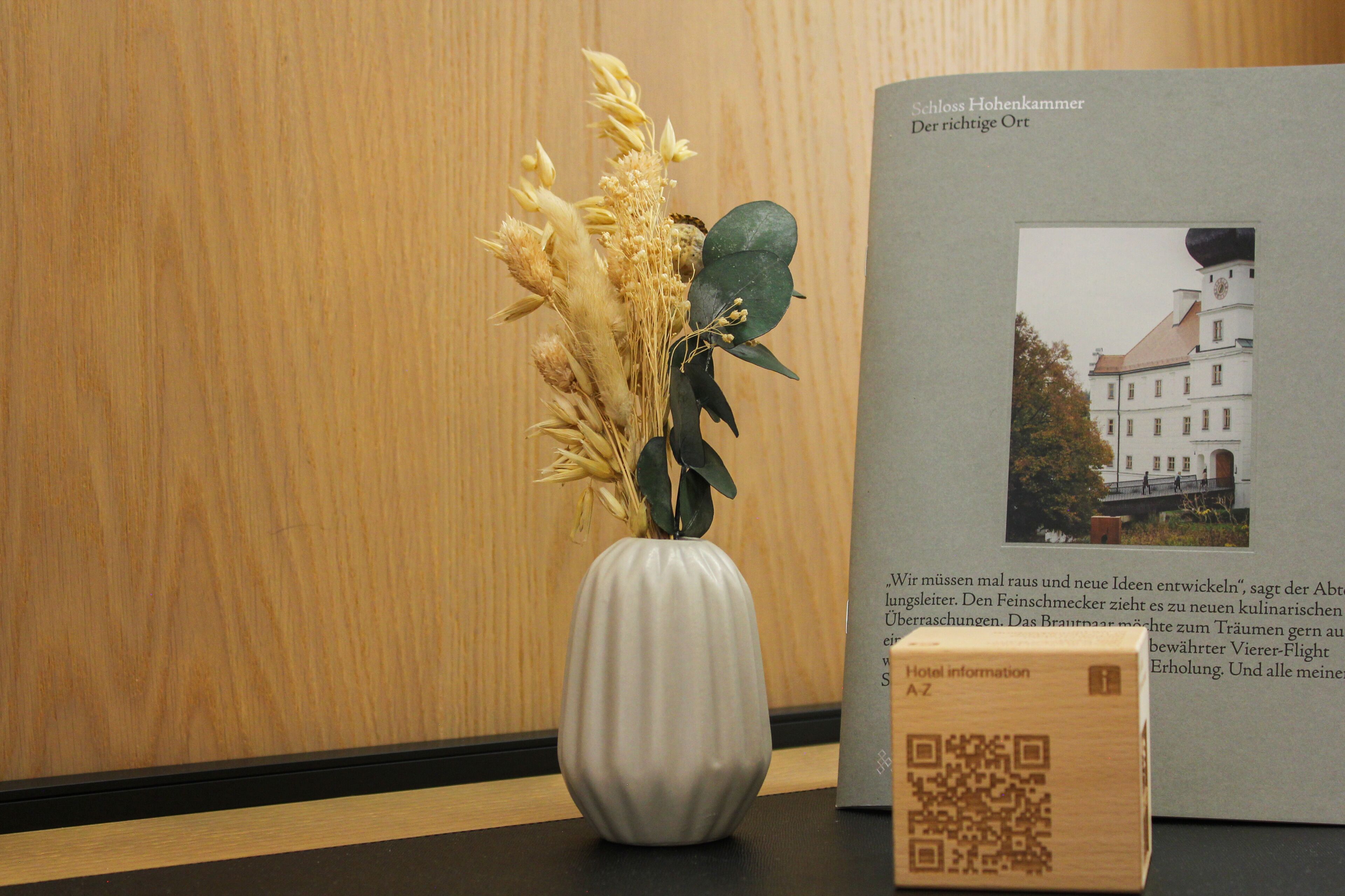 Un jarrón blanco con flores secas, un folleto de hotel impreso y un código QR sobre una mesa.