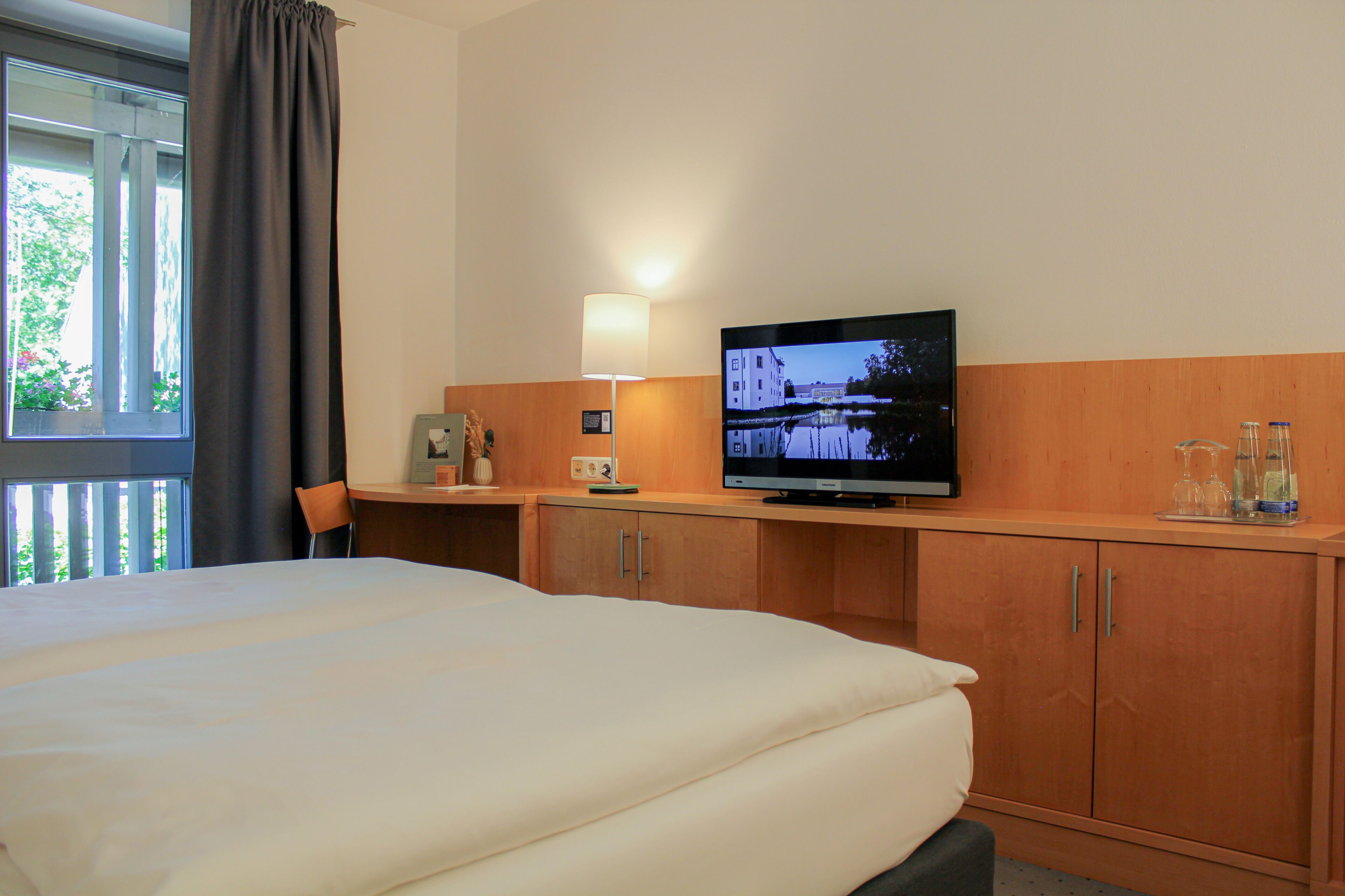 Una habitación de hotel con una cama, televisión, escritorio, silla y ventana con cortinas.
