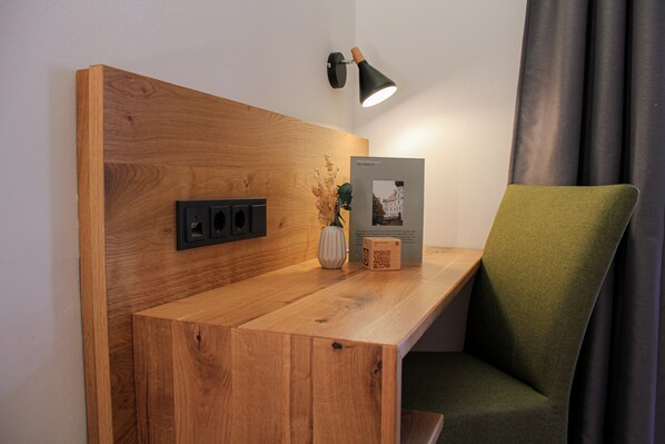 A wooden desk with a green chair, a wall-mounted lamp, a vase with dried flowers, and a framed picture.
