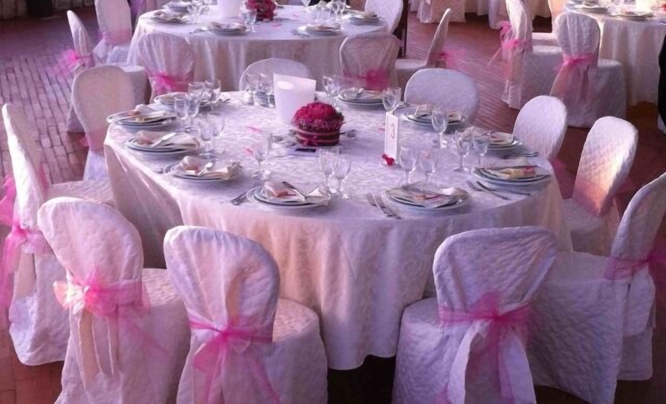 Round tables with white tablecloths and chairs with pink sashes, set for a formal event.