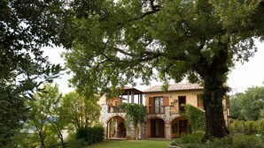 A stone house with a balcony, arched windows, and a large tree in the foreground.