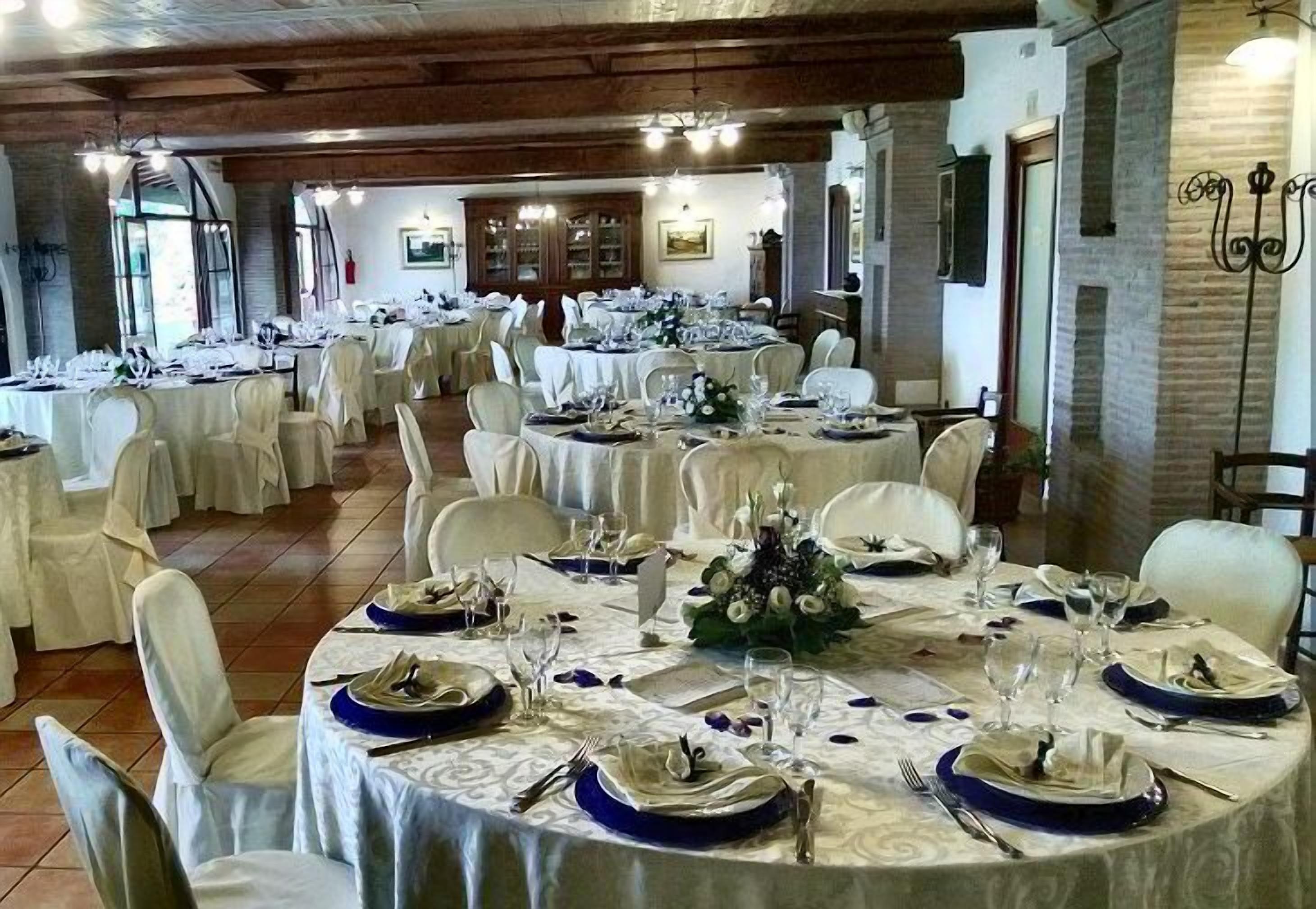 A banquet hall with round tables set for a formal event, featuring white and blue tablecloths, and a rustic wooden ceiling.