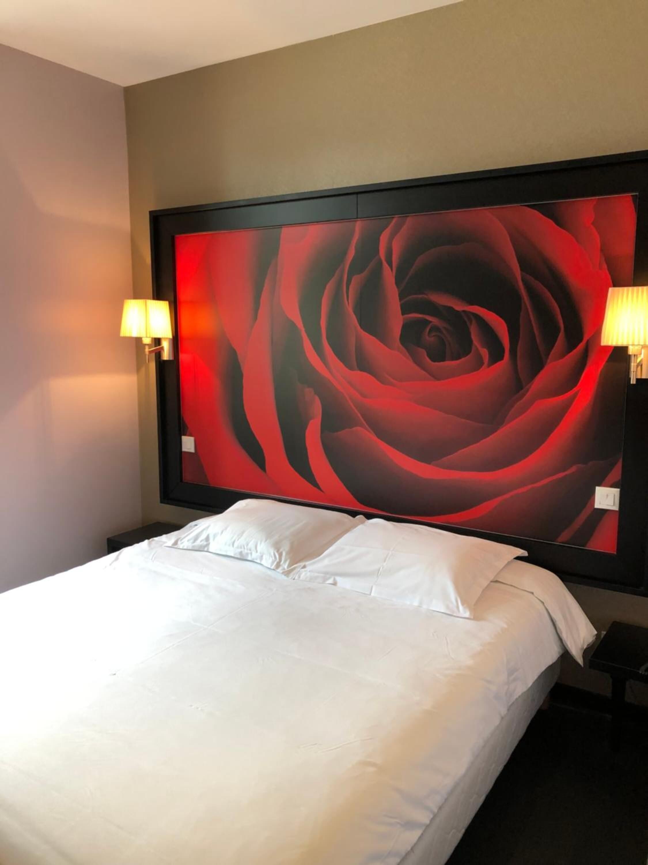 A hotel room with a large wall mural of a red rose, a white bed, and two wall-mounted lamps.