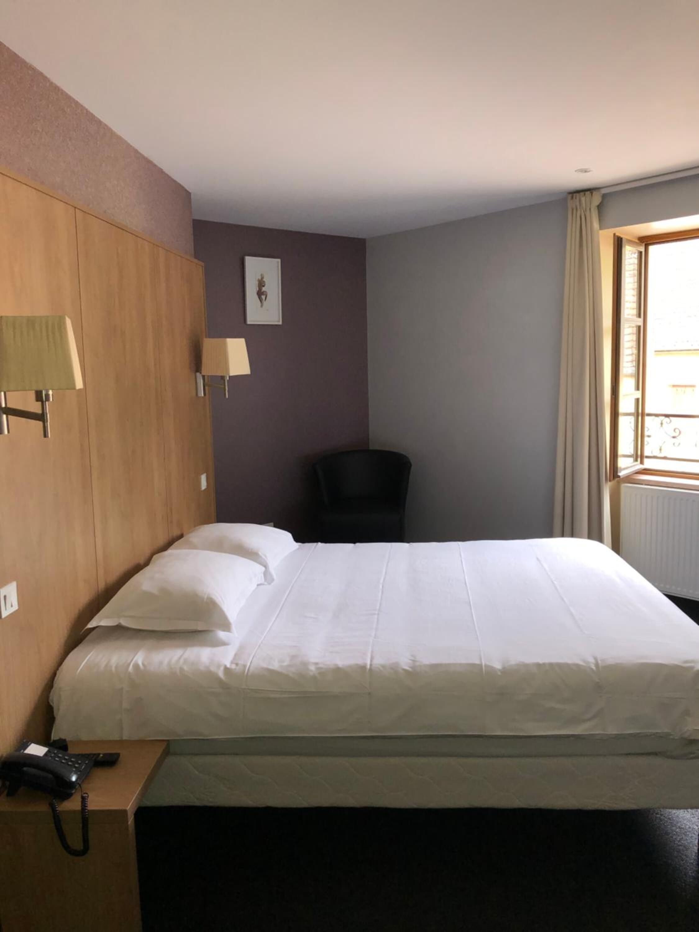 Habitación de hotel con una cama, una silla, un escritorio con teléfono y una ventana con cortinas.