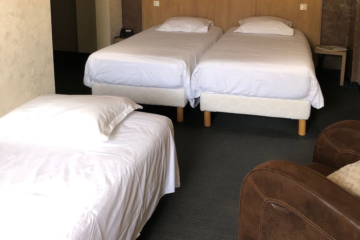 A hotel room with two single beds, a brown leather armchair, and a wooden wardrobe with two wall-mounted lamps.