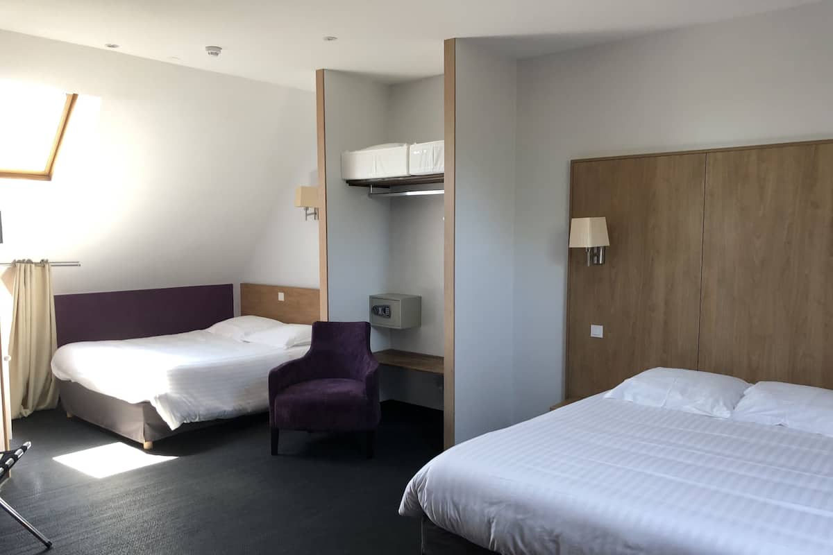 A hotel room with two beds, a purple armchair, a desk with a lamp, and a wardrobe.