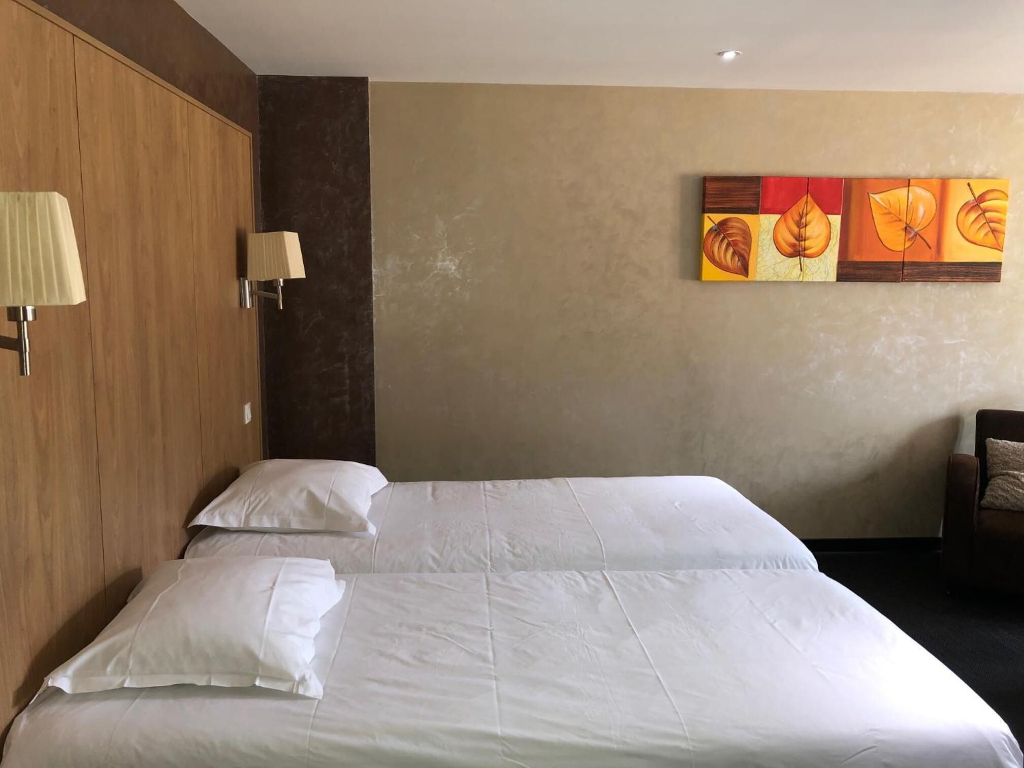 A hotel room with two beds, a wooden wardrobe, a wall-mounted lamp, and a painting of leaves.