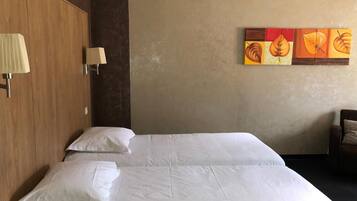 A hotel room with two beds, a wooden wardrobe, a wall-mounted lamp, and a painting of leaves.