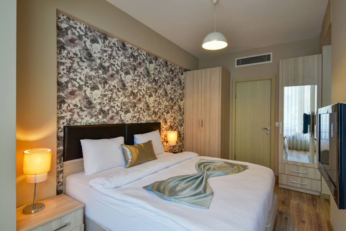 Un lit bien fait, agrémenté d’un ruban décoratif, une table de chevet avec une lampe, une armoire et un miroir.