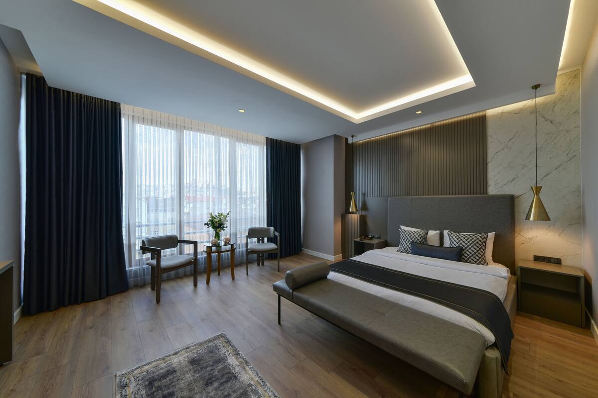 Une chambre d’hôtel moderne dotée d’un grand lit, de tables de chevet, d’un coin salon avec des fauteuils et offrant une vue sur la ville grâce à de grandes fenêtres.