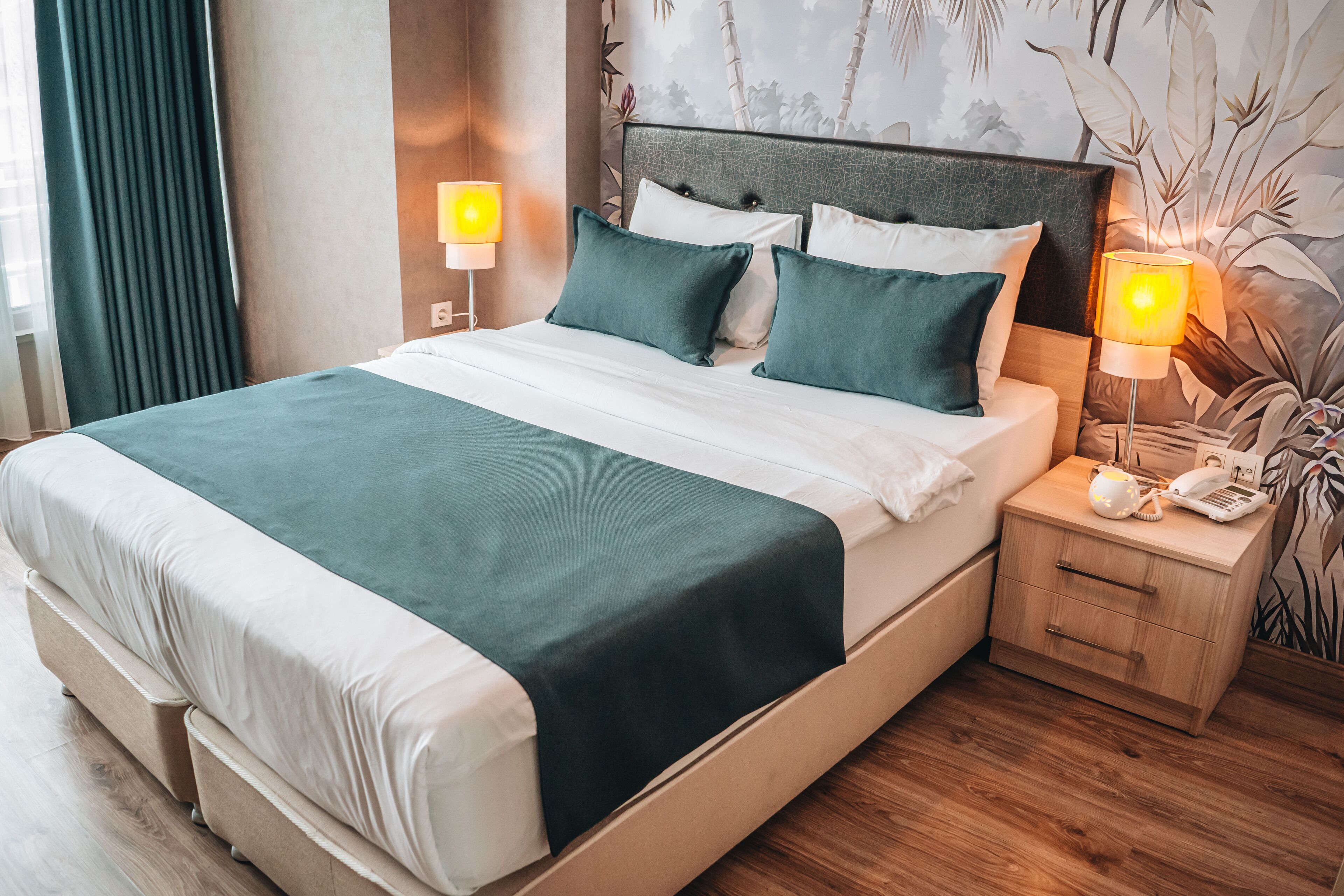 Una cama bien hecha con colcha verde y ropa blanca, una mesita de noche de madera con lámpara, y una pared con un mural de temática natural.