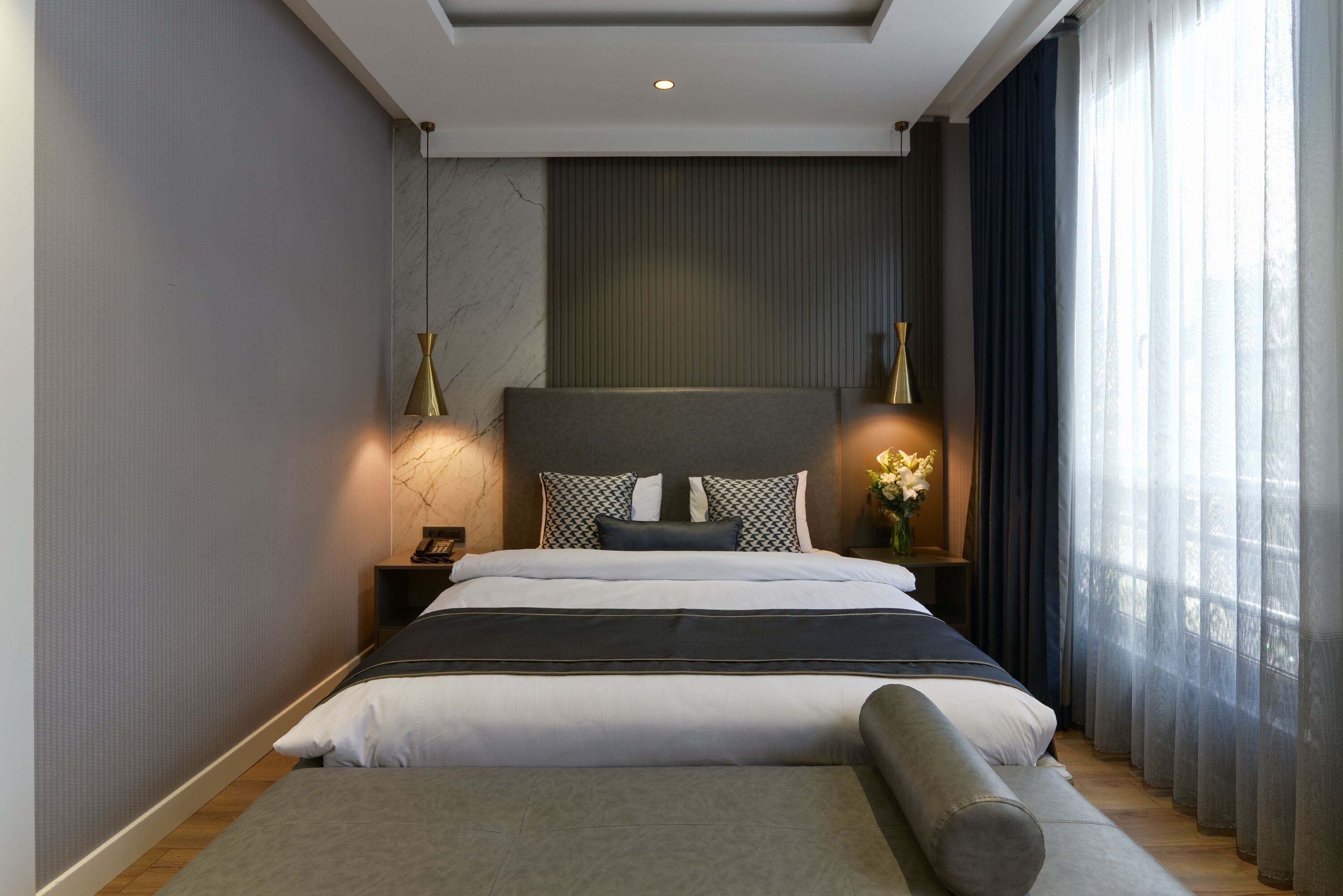 A modern bedroom with a large bed, a headboard, bedside lamps, and a view of the outdoors through sheer curtains.