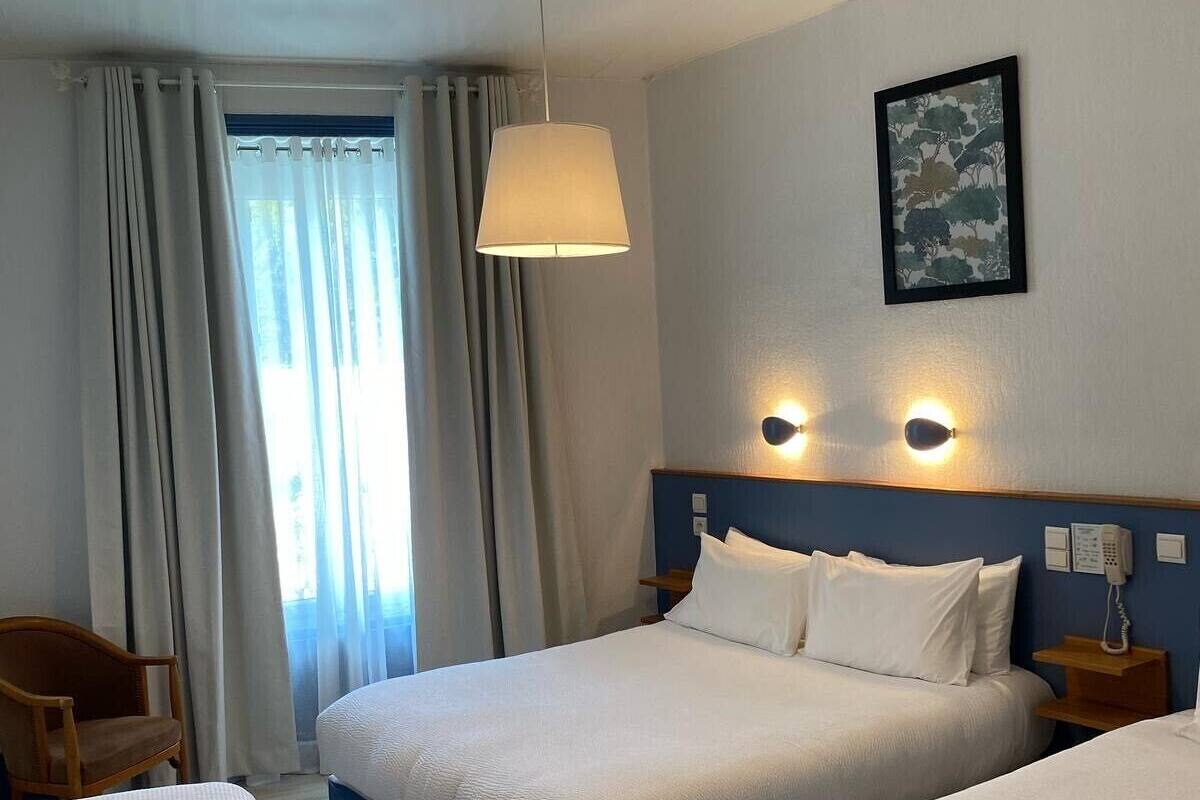 A hotel room with two beds, a chair, a window with curtains, a hanging lamp, a wall-mounted phone, and a framed picture.