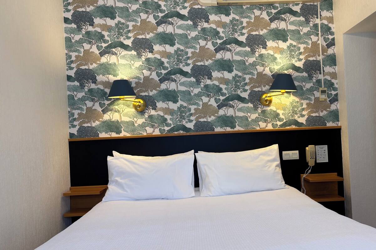 A hotel room with a bed, two wall-mounted lamps, a telephone, and a wall with a tree pattern.
