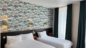 Habitación de hotel con dos camas, papel tapiz estampado, una mesita de noche con lámpara y una ventana con cortinas.