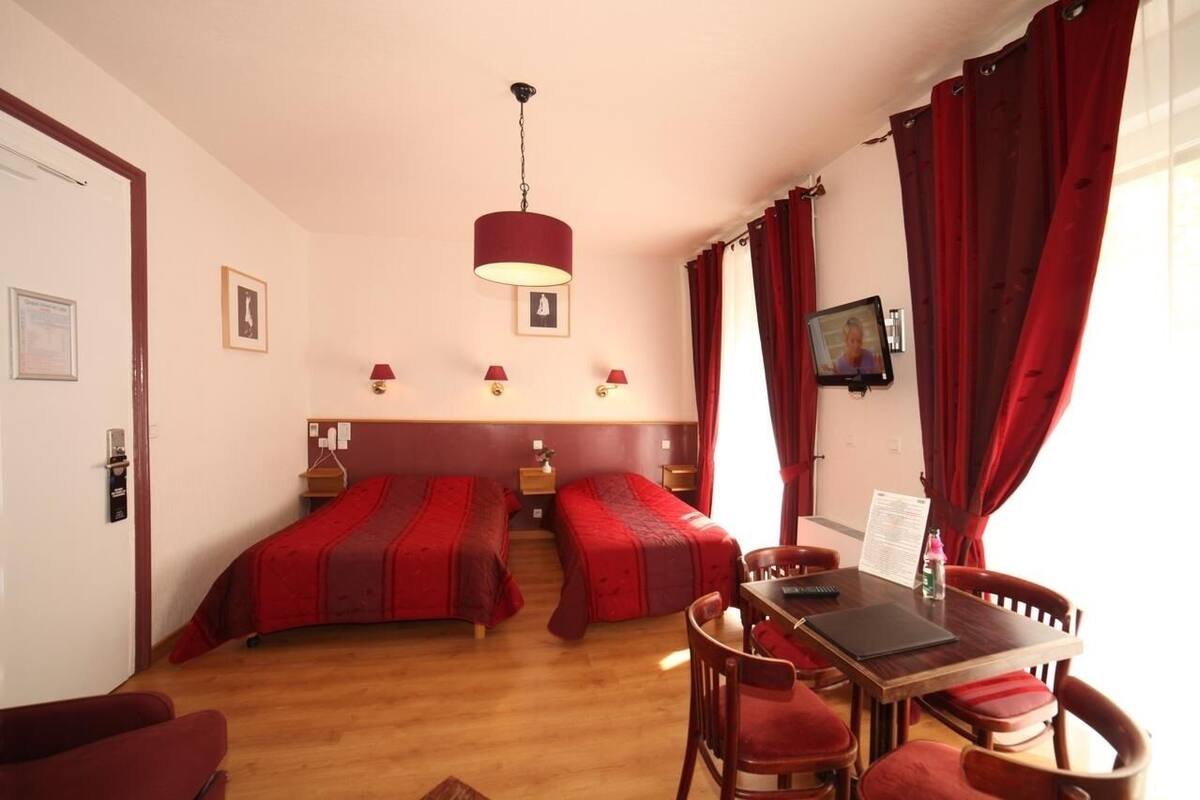 A hotel room with two single beds, a dining table with chairs, a television, and a window with red curtains.