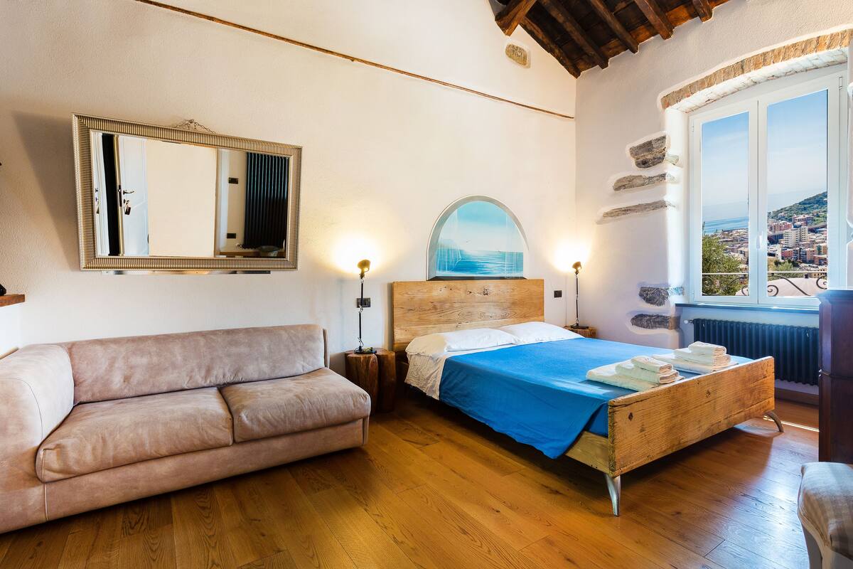 A bedroom with a wooden bed, a sofa, a large mirror, and a view of a cityscape through a window.