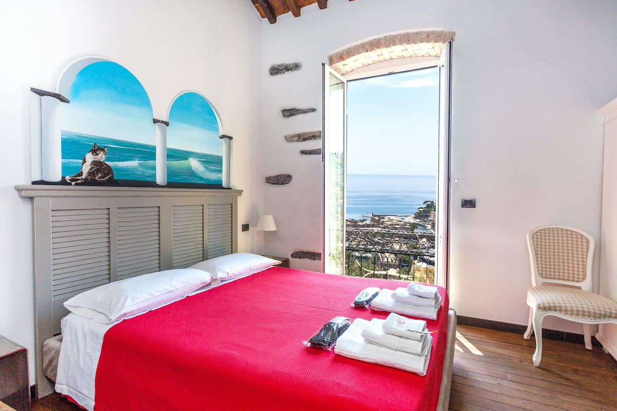 A hotel room with a bed, a chair, a window with a view of the sea, and a painting of a cat on the wall.