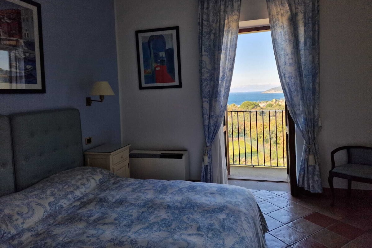 A bedroom with a bed, a nightstand, a lamp, a chair, and a balcony with curtains.