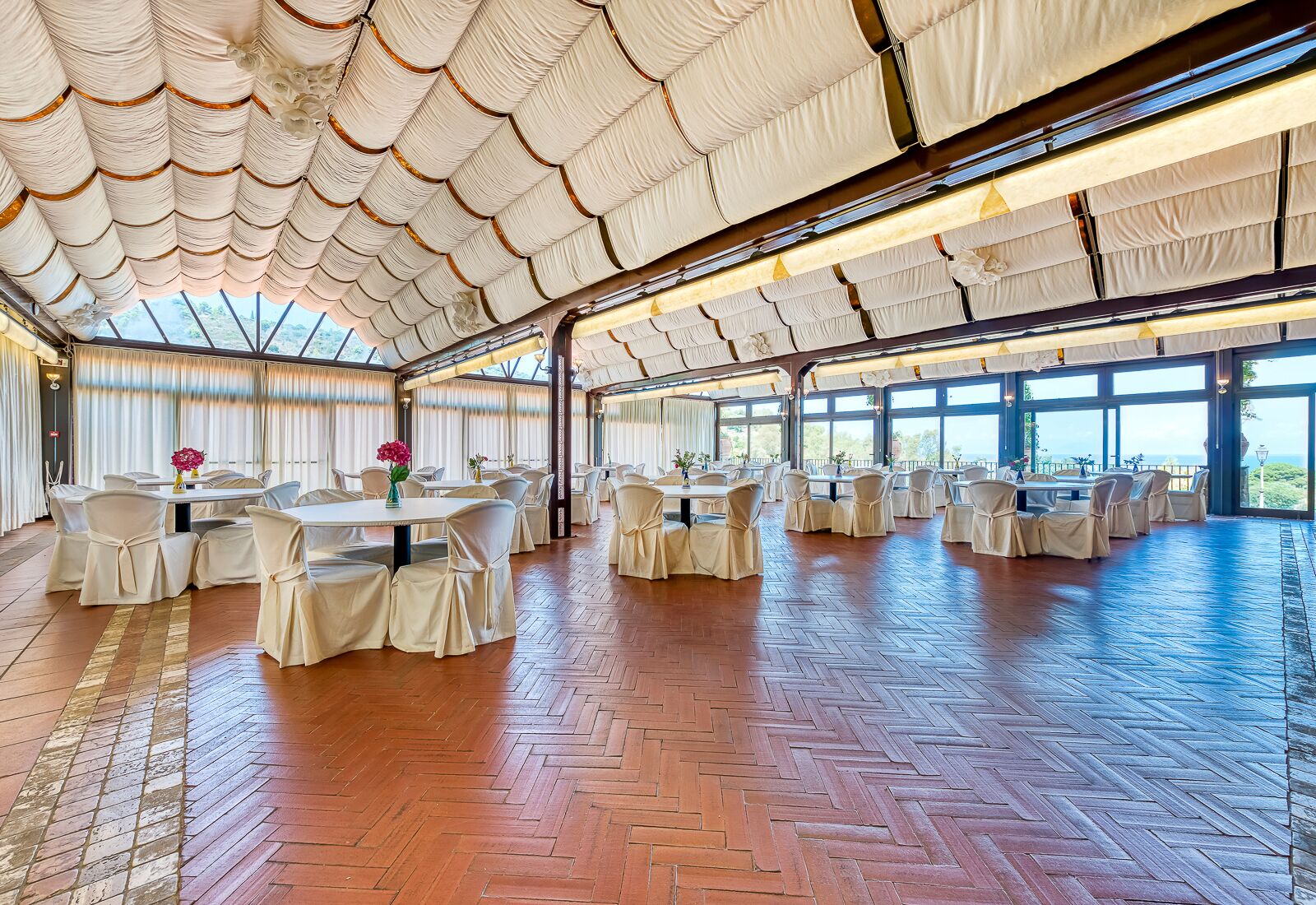 A spacious indoor venue with multiple round tables and chairs, large windows, and a wooden herringbone floor.